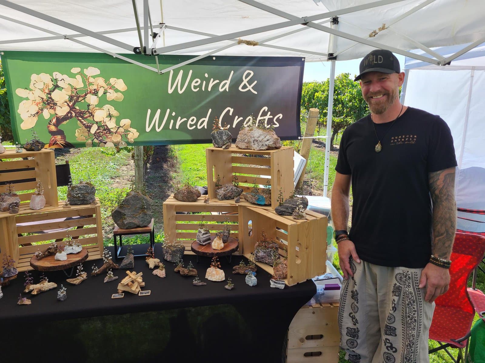 Matt Quast of Weird & Wired Crafts poses with his nature-inspired crystal and wire art, including trees and landscapes, at the outdoor market. 