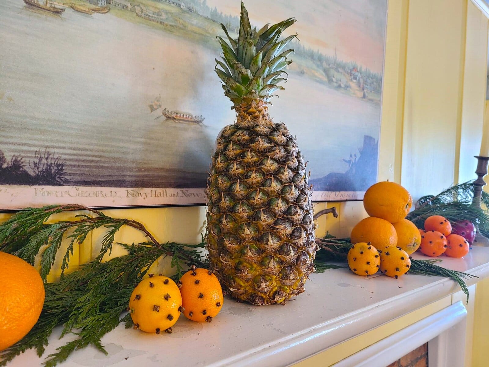 A pineapple anchors a mantel display surrounded by citrus fruit pomanders and evergreen bundles. Guides explained that pineapples were rare imported items in the early-1800s and signalled status and abundance. 