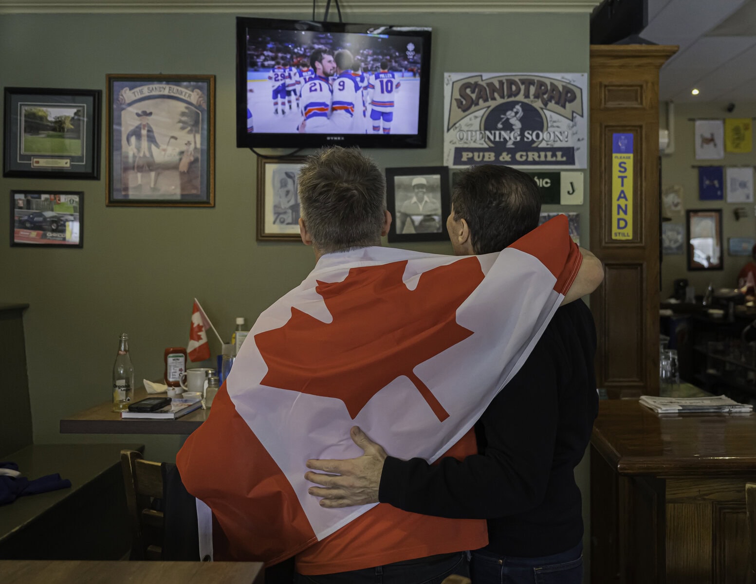 As Team U.S.A. celebrates its gold-medal win, scoring the winning goal in overtime, two Sandtrap patrons console themselves after another Olympic loss for Team Canada.