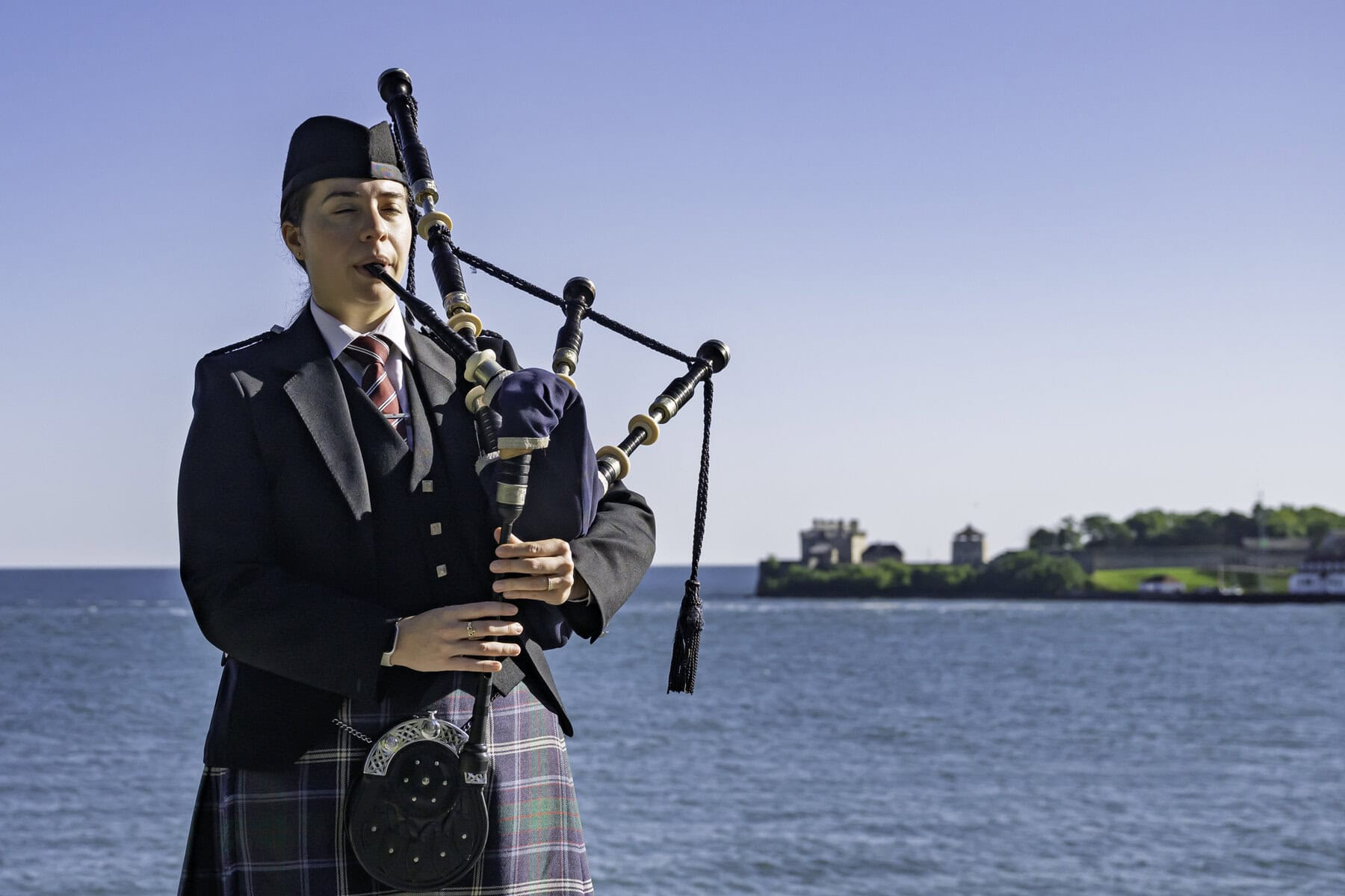 Victoria McRae plays the bagpipes at the shores of the Niagara River — visible in the background is the United States itself, and Fort Niagara.