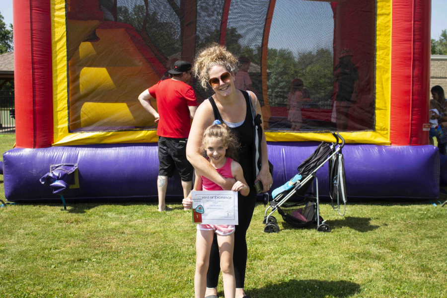 Tennille Chwalczuk and Daughter Mia holding a Service of Excellence Award from the Niagara-on-the-Lake Skating Club, in front of the Bouncy Castle at the Community Centre Open-House